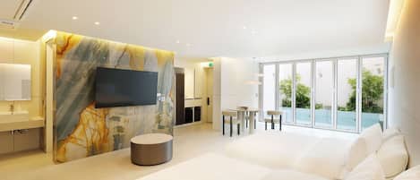 A modern hotel room with a large bed, a flat-screen TV, a marble accent wall, a dining area with a table and chairs, and a balcony with plants.