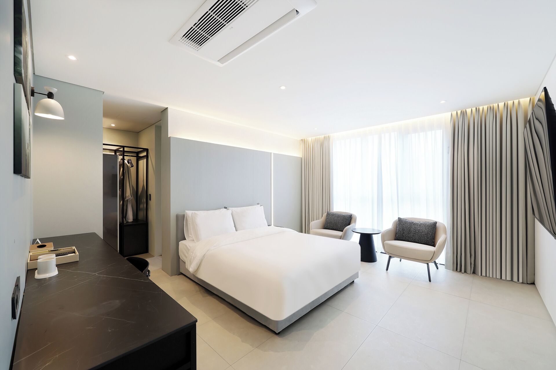 A modern hotel room with a large bed, a desk, two armchairs, and a view of the city.