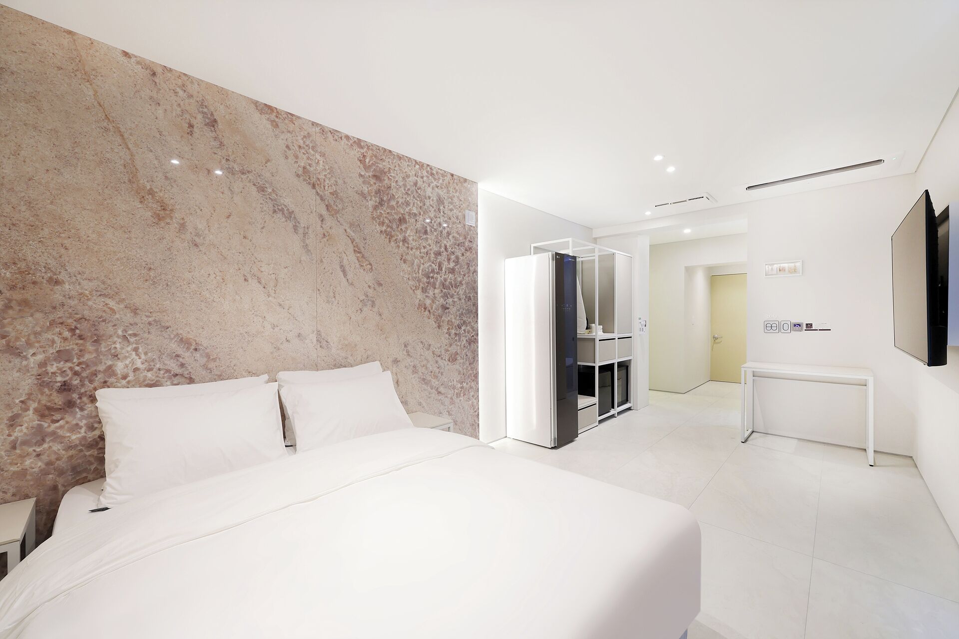 Una habitación de hotel moderna con una cama grande, un detalles en pared de mármol, un minibar y una televisión.