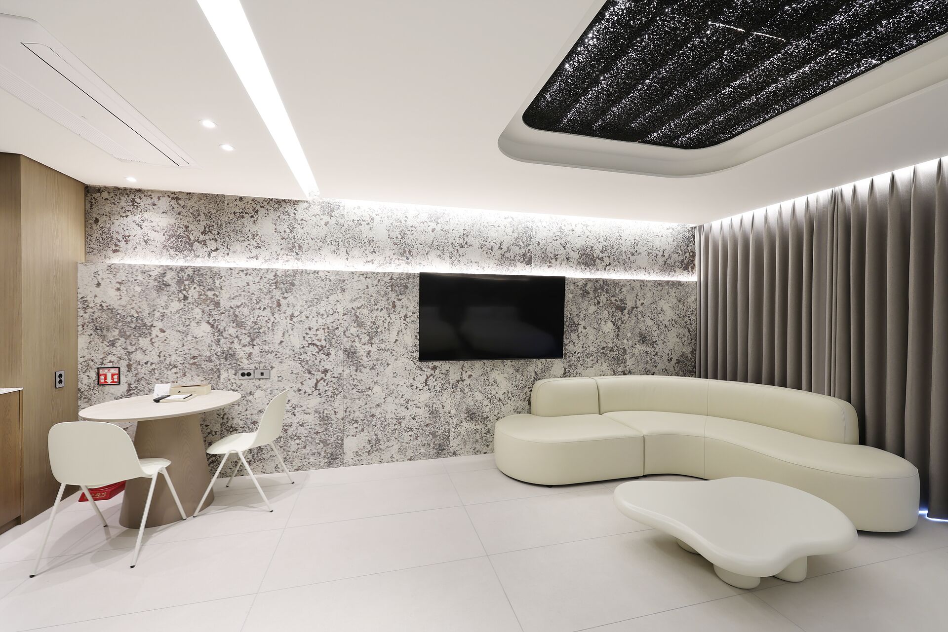 Un salon moderne avec un canapé d’angle, un téléviseur à écran plat fixé au mur, une table de salle à manger ronde avec des chaises, et un plafond au motif texturé.