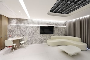 A modern living room with a sectional sofa, a flat-screen TV mounted on the wall, a round dining table with chairs, and a ceiling with a textured design.