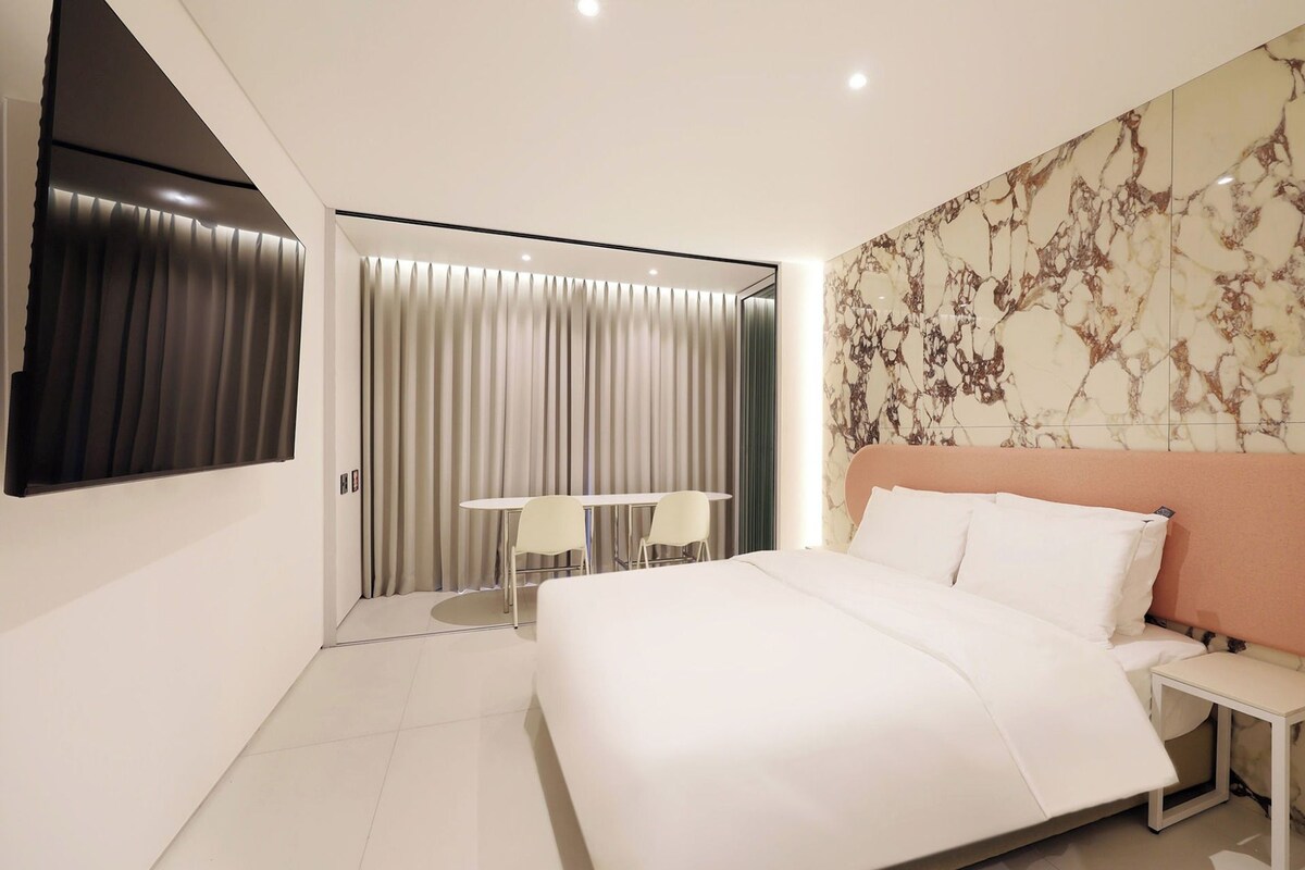 A modern bedroom with a large bed, a flat-screen TV mounted on the wall, a marble-patterned wall, and a sliding door leading to a balcony.