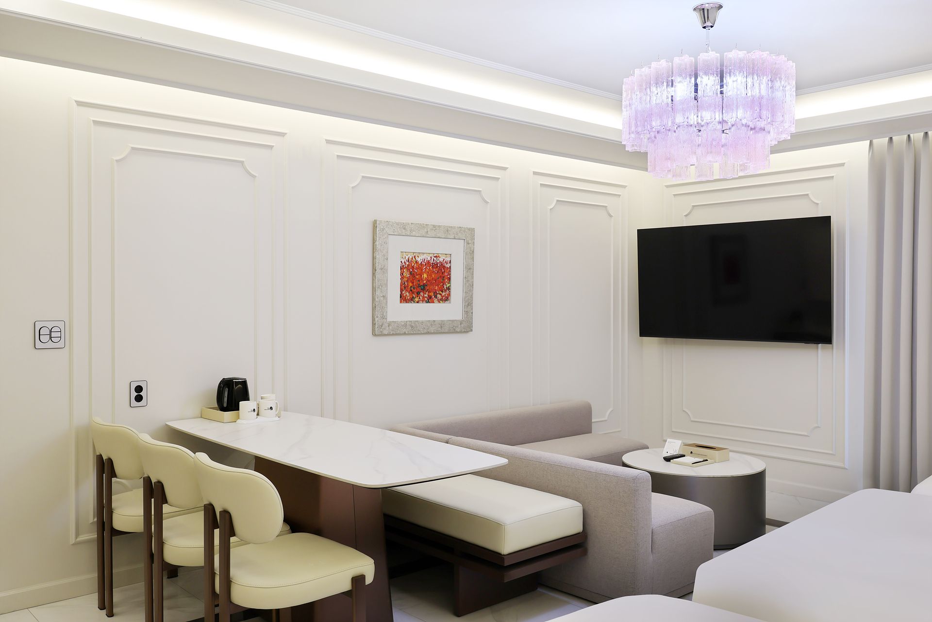 A modern hotel room with a dining area, a sofa, a small round table, a television mounted on the wall, and a chandelier.