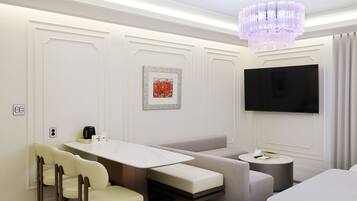 A modern hotel room with a dining area, a sofa, a small round table, a television mounted on the wall, and a chandelier.