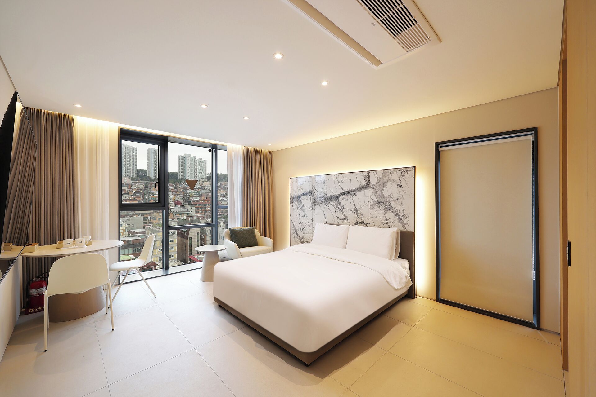 A hotel room with a bed, a desk, a chair, and a view of the city.