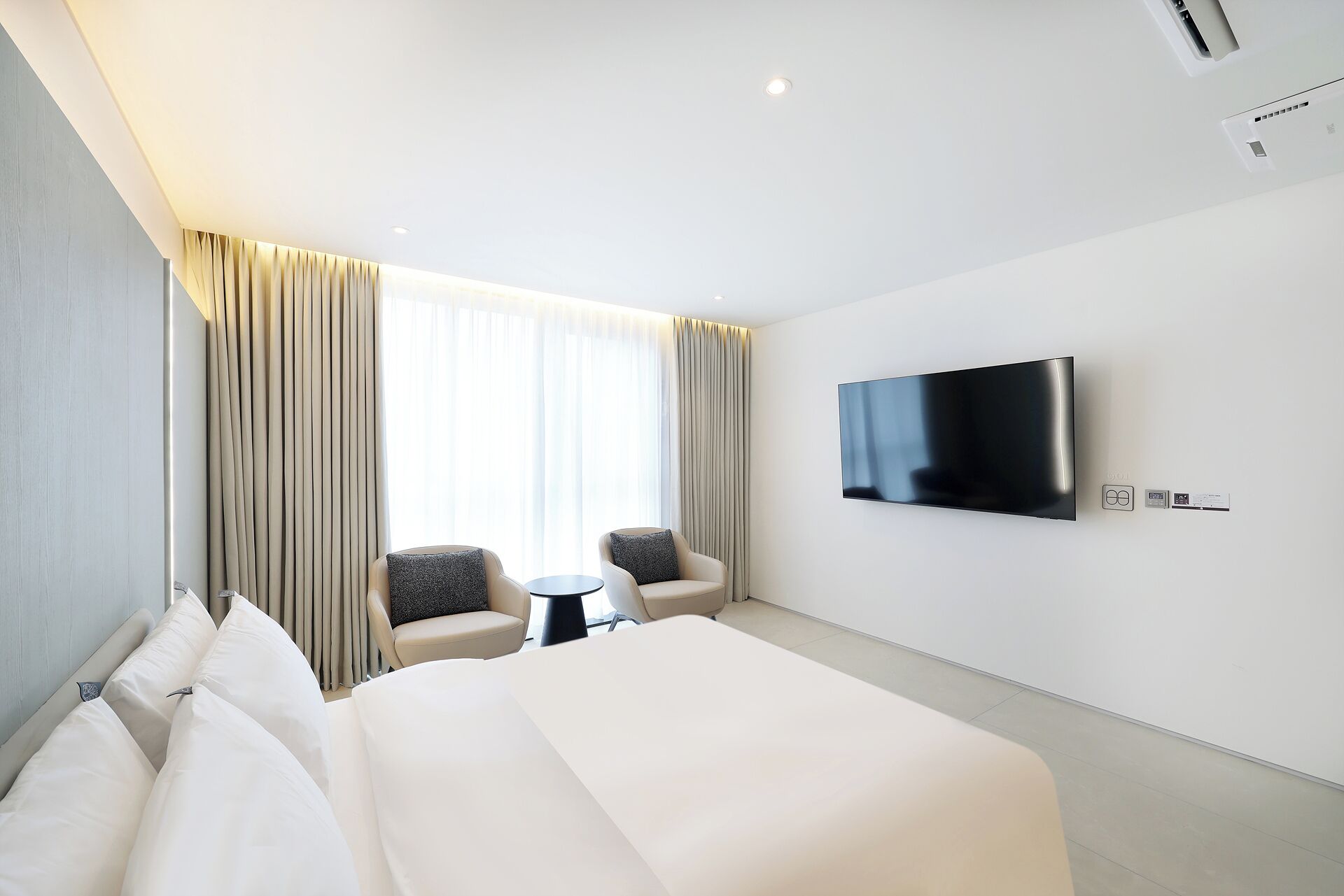 A hotel room with a large bed, two armchairs, a small table, a flat-screen TV mounted on the wall, and a window with curtains.
