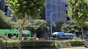 A modern building with a reflective glass facade, surrounded by trees and a street with vehicles.