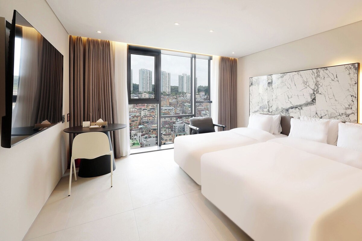A hotel room with a large bed, a desk, a chair, a TV, and a city view.