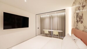 A modern hotel room with a large bed, a flat-screen TV mounted on the wall, a small round table with two chairs, and a curtain-covered window.
