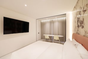 Una habitación de hotel moderna con una cama grande, un televisor de pantalla plana en la pared, una mesita redonda con dos sillas y una ventana con cortinas.