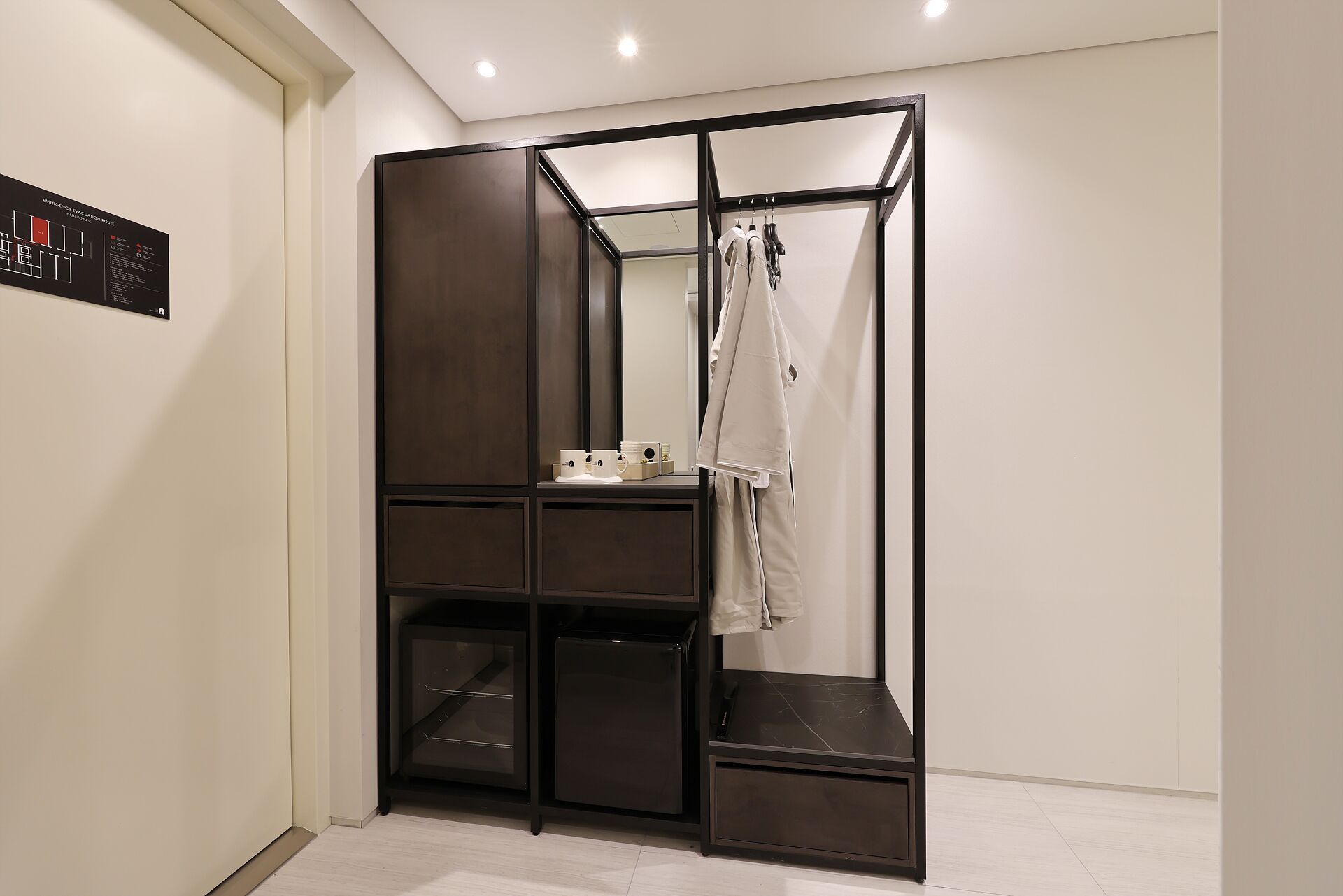 Ein kleines Zimmer mit Kleiderschrank, einem Tisch mit Tasse und einer Kleiderstange mit Kleidung.