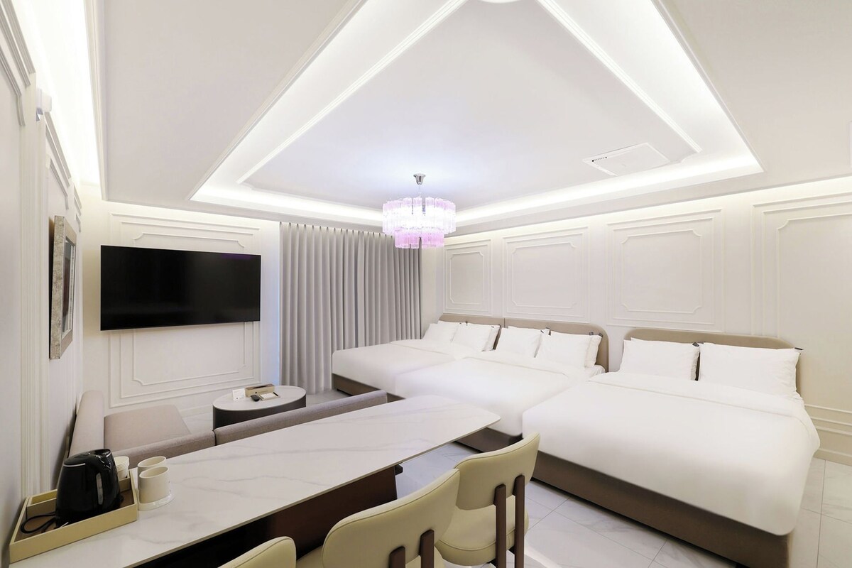 A hotel room with a flat-screen TV mounted on the wall, a bed with white bedding, a small round table with a kettle and cups, and a chandelier.
