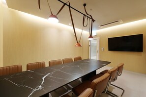 Un salón de conferencias con una mesa larga, sillas de cuero marrón, iluminación colgante, un equipo de aire acondicionado y un televisor de pantalla plana en la pared.