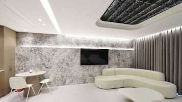 Una sala moderna con un sofá curvo, un televisor de pantalla plana montado en la pared, una mesa redonda con sillas y una lámpara de techo.