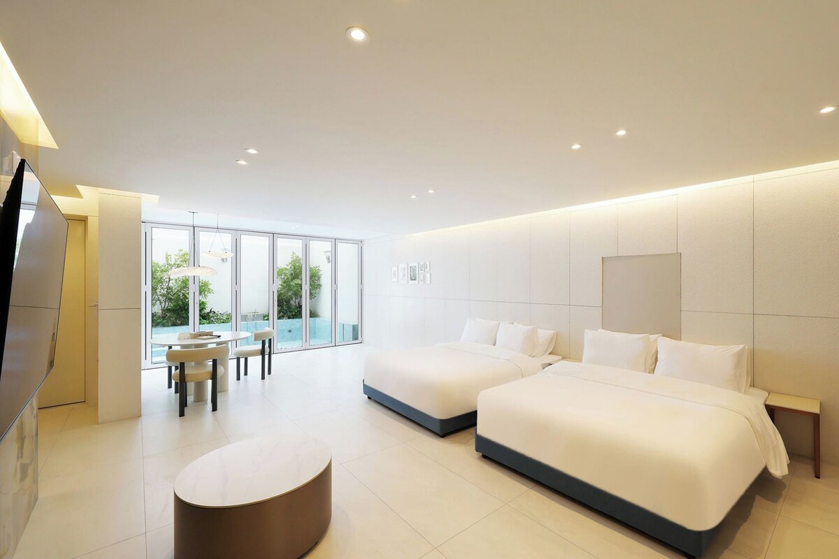 A modern hotel room with two beds, a dining area, and a large window.