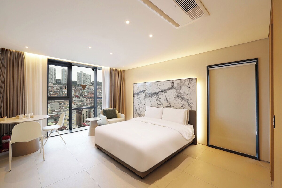 A modern hotel room with a large bed, a desk, and a chair, a city view through the window, and a marble headboard.
