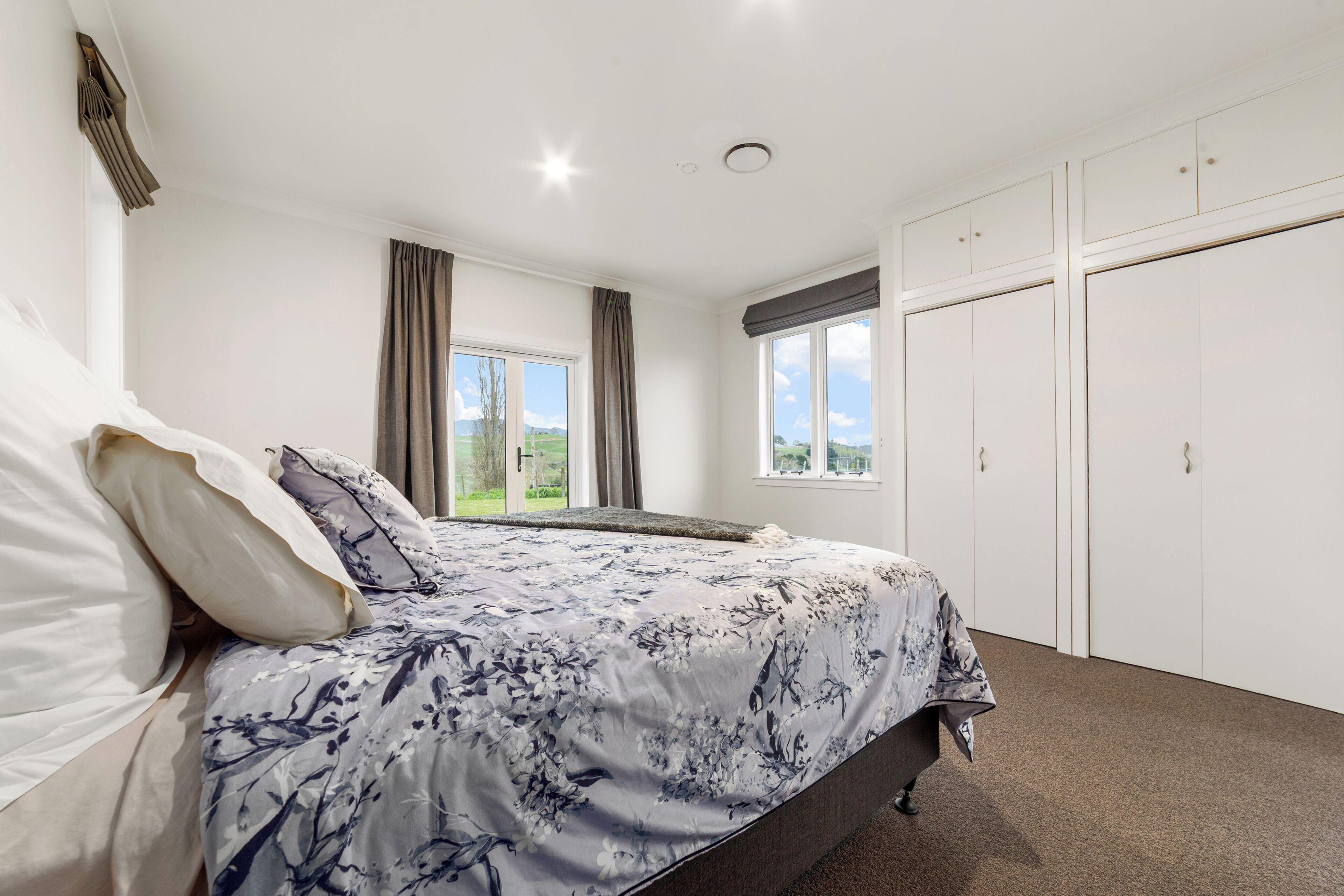 A bedroom with a bed, two windows with curtains, a wardrobe, and a view of the outdoors.
