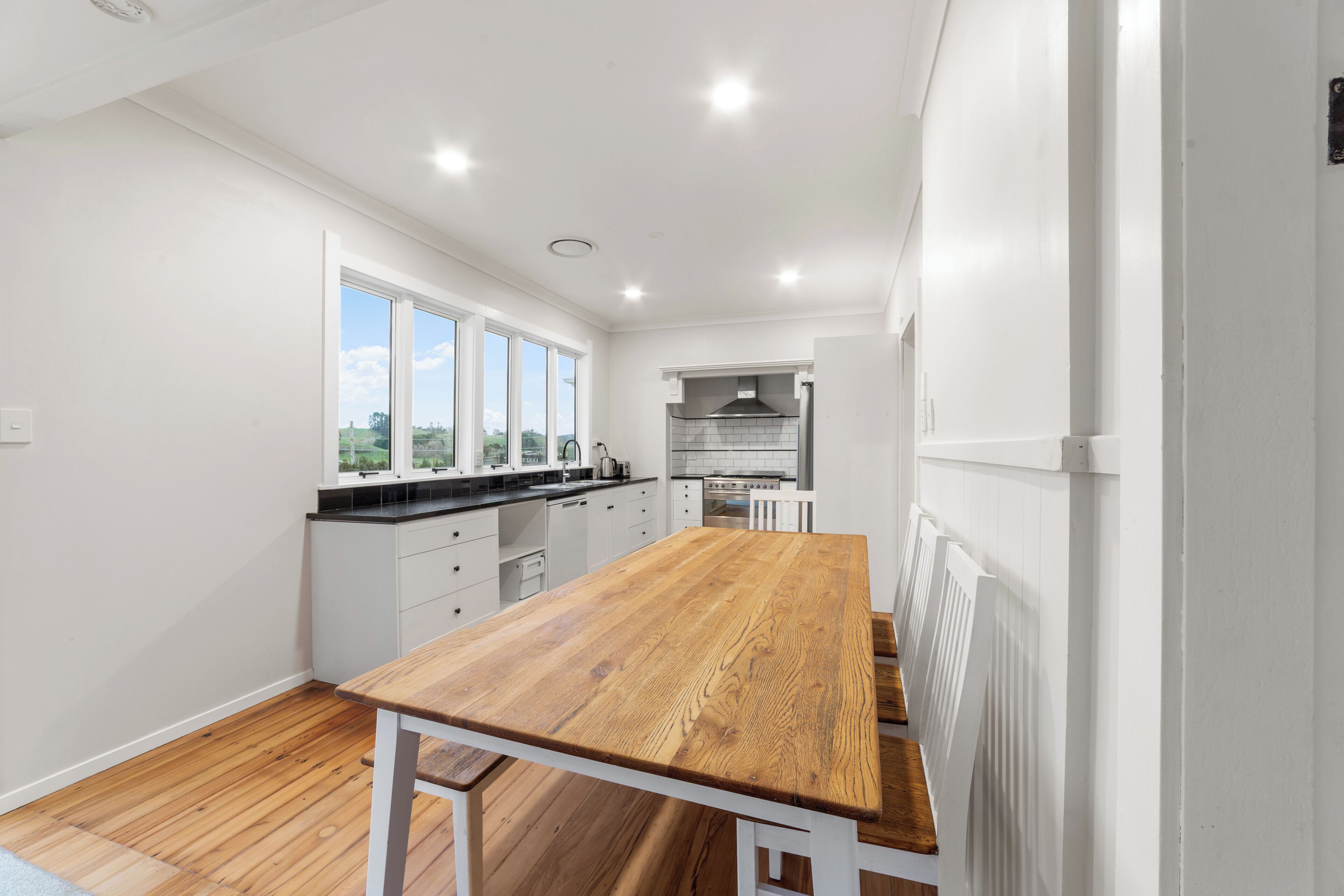 A modern kitchen with white cabinets, a wooden table, and a view of the countryside through large windows.