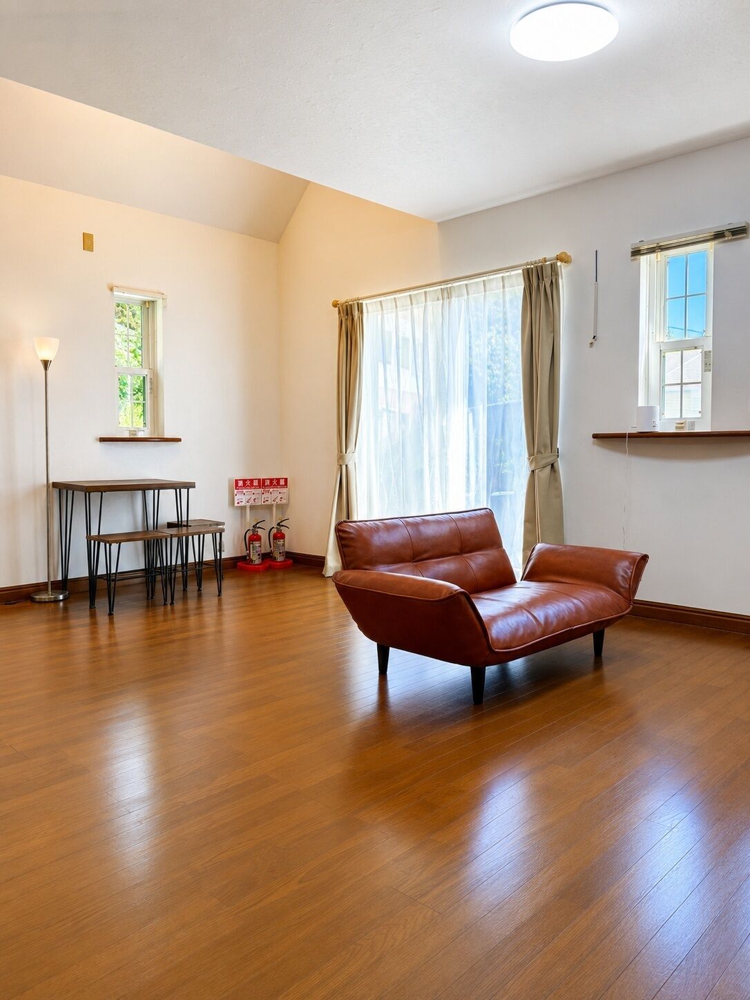 A spacious room with a leather sofa, a small table, and a chair. There are two windows with curtains, a ceiling light, and a fire extinguisher on the floor.