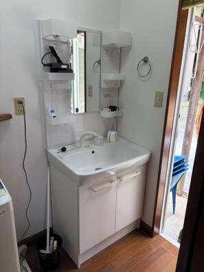 Un baño con lavamanos blanco, espejo y armarios para guardar objetos. Se ve un trapeador y un cubo, además de un enchufe en la pared.