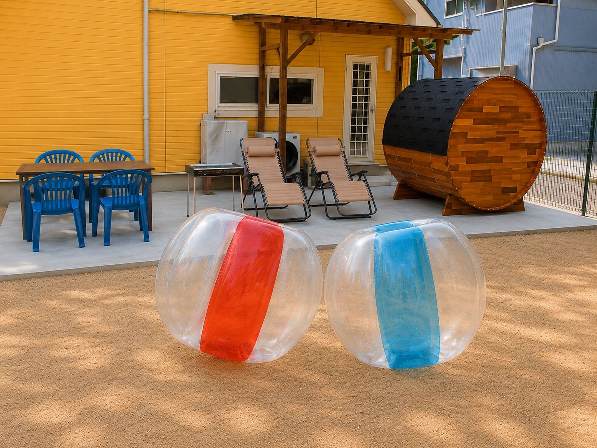 Two transparent inflatable balls, a wooden barrel, and outdoor furniture against a yellow building.