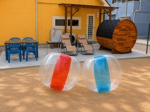 Dos bolas inflables transparentes, un barril de madera y mobiliario exterior frente a un edificio amarillo.