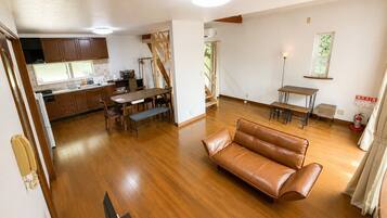 A spacious living area with a leather sofa, wooden flooring, a dining table with chairs, a kitchen area with wooden cabinets, and a staircase leading to another level.
