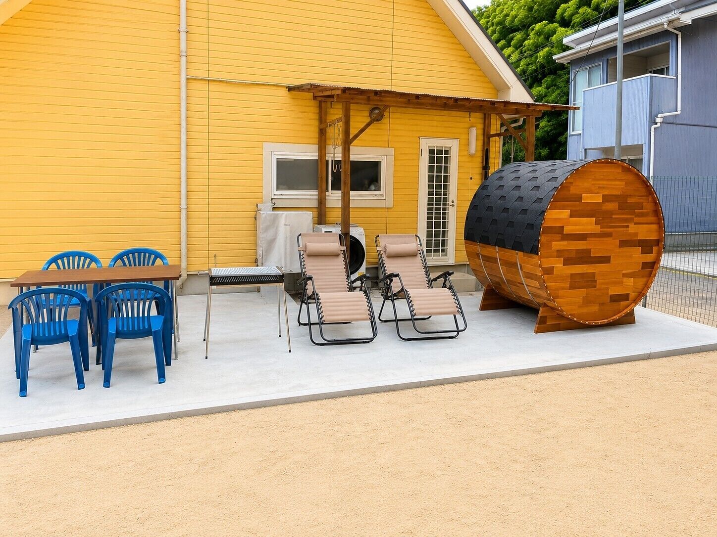 A yellow building with a covered patio area featuring a table, chairs, and a large barrel.