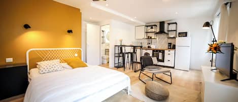 A modern bedroom with a bed, a small seating area, and a kitchenette in the background.
