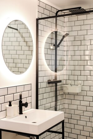 A modern bathroom with a white sink, a round mirror, and a glass-enclosed shower. - Sunshine Apartment - Ancient Theater 30 meters away (Orange)