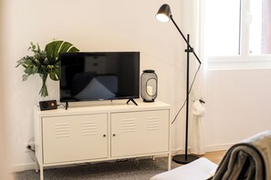 Ein Zimmer mit einem Flachbildfernseher auf einer weißen Kommode, einer Stehlampe, einem Blumentopf und einem Fenster mit Vorhängen.