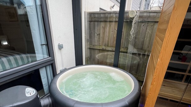 A hot tub on a wooden deck with a glass enclosure, a control panel, and a wooden ladder.