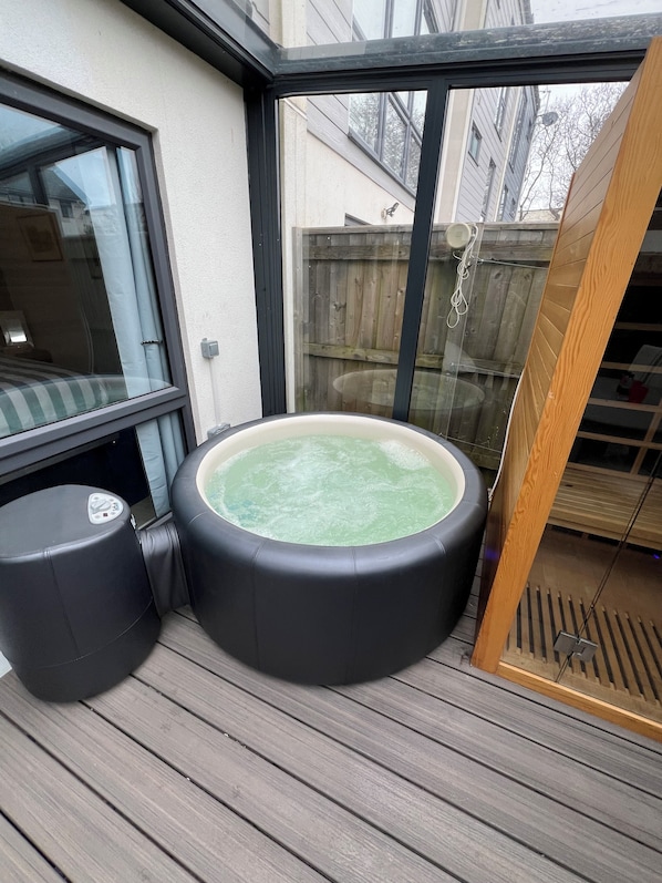 A hot tub on a wooden deck with a glass enclosure, a control panel, and a wooden ladder.