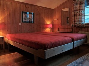 Ein Zimmer mit Holzwänden, einem Bett, einem Nachttisch, einer Lampe und einem mit Vorhängen versehenen Fenster.