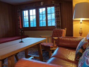Ein gemütliches Zimmer mit einem Holztisch, einer Couch und einem Sessel. Rechts ist ein Fenster mit Vorhängen und eine Lampe zu sehen.