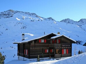 Eine Holzhütte mit roten Fensterläden vor einer verschneiten Berglandschaft.
