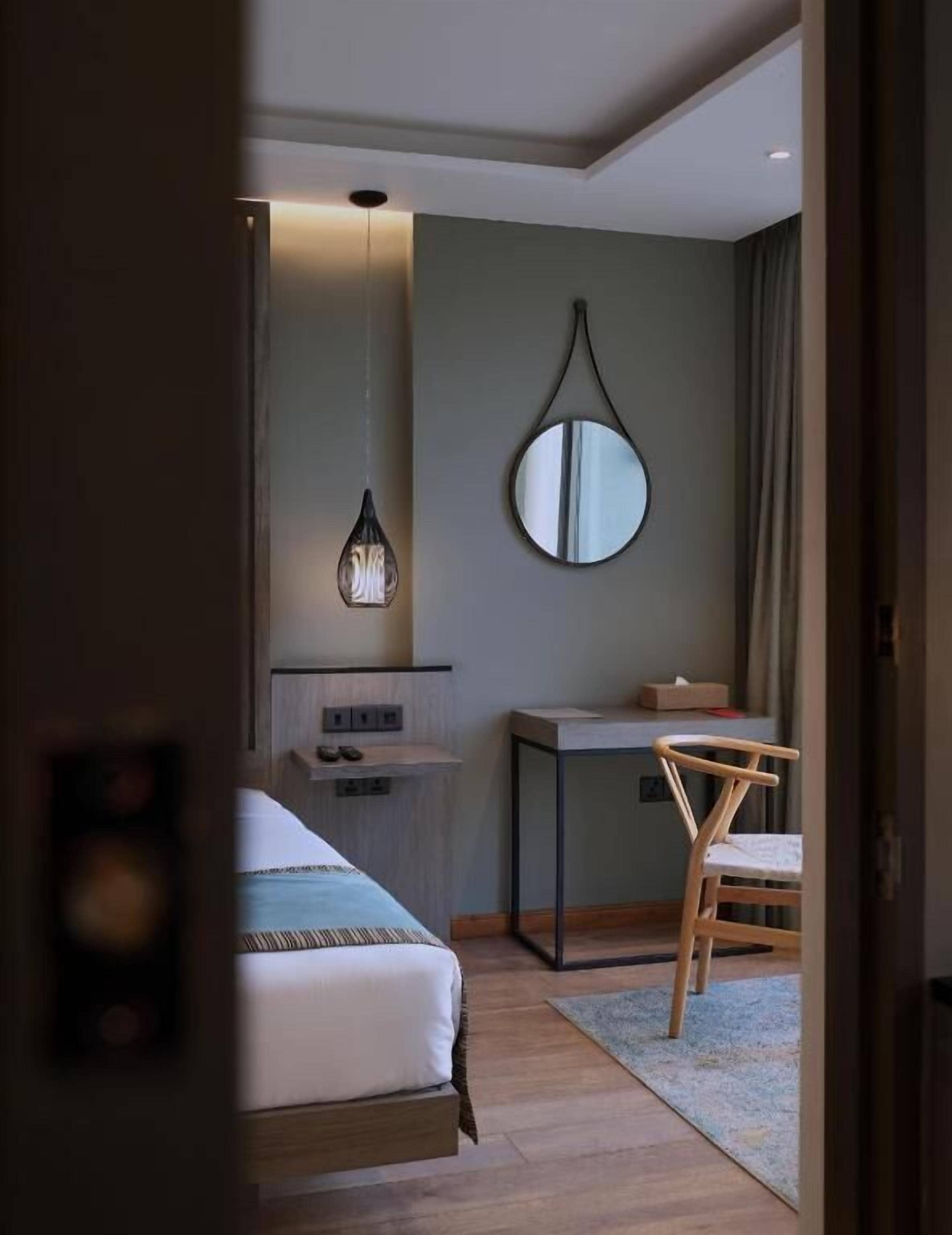 Ett hotellrum med en säng, ett skrivbord, en stol, en spegel och en hängande lampa.