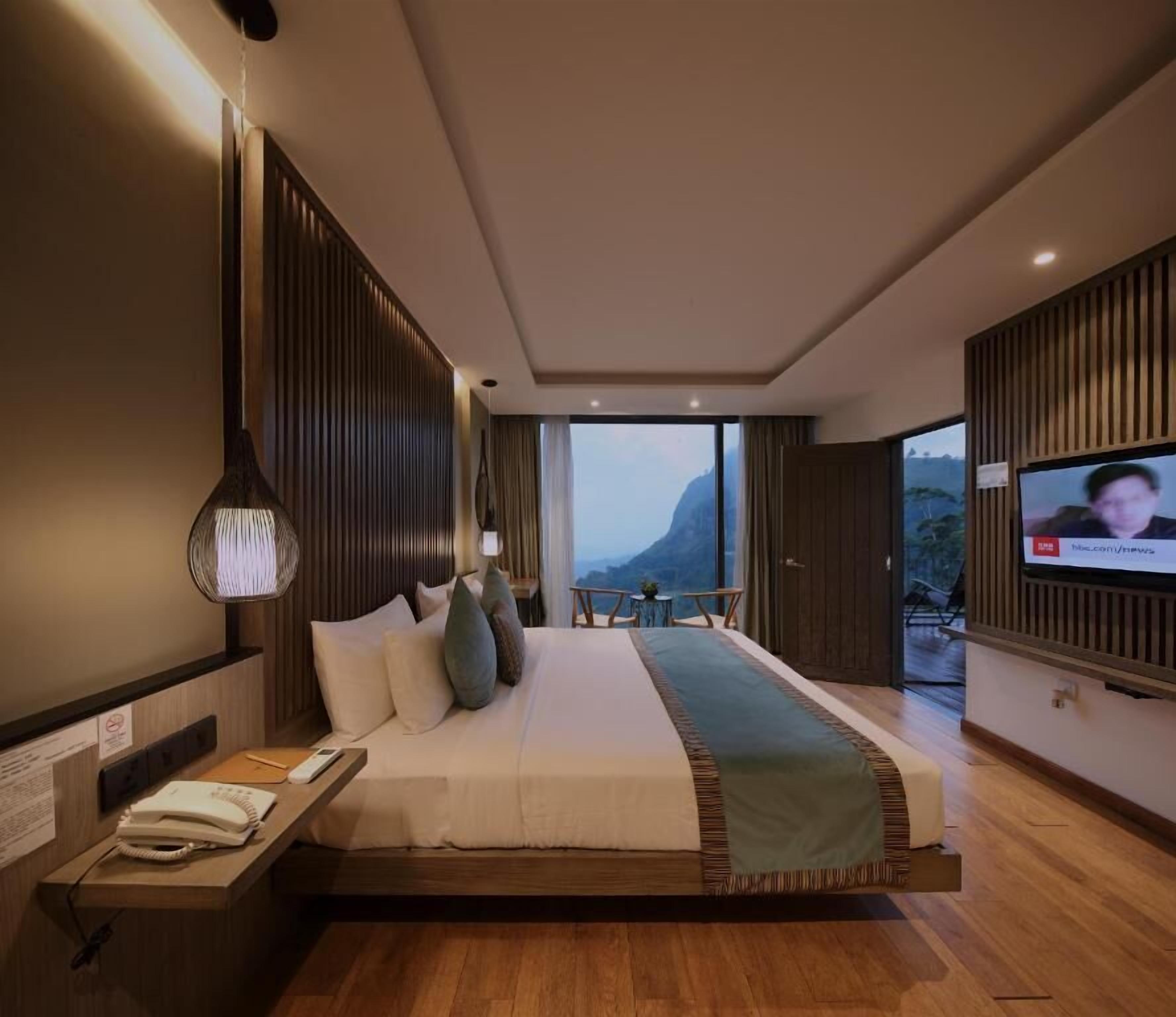Ein modernes Hotelzimmer mit einem großen Bett, einem Fernseher, einem Schreibtisch mit Telefon und Blick ins Freie.