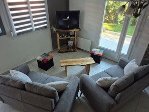 A living room with two grey sofas, a wooden coffee table, a television on a stand, and a window with blinds. - A 3-star vacation home with great amenities. (Le Tholy)