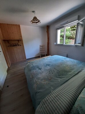 A bedroom with a bed, a wooden wardrobe, a chair, a window with a view, and a hanging light fixture. - Renovated house near Gérardmer (Le Tholy)
