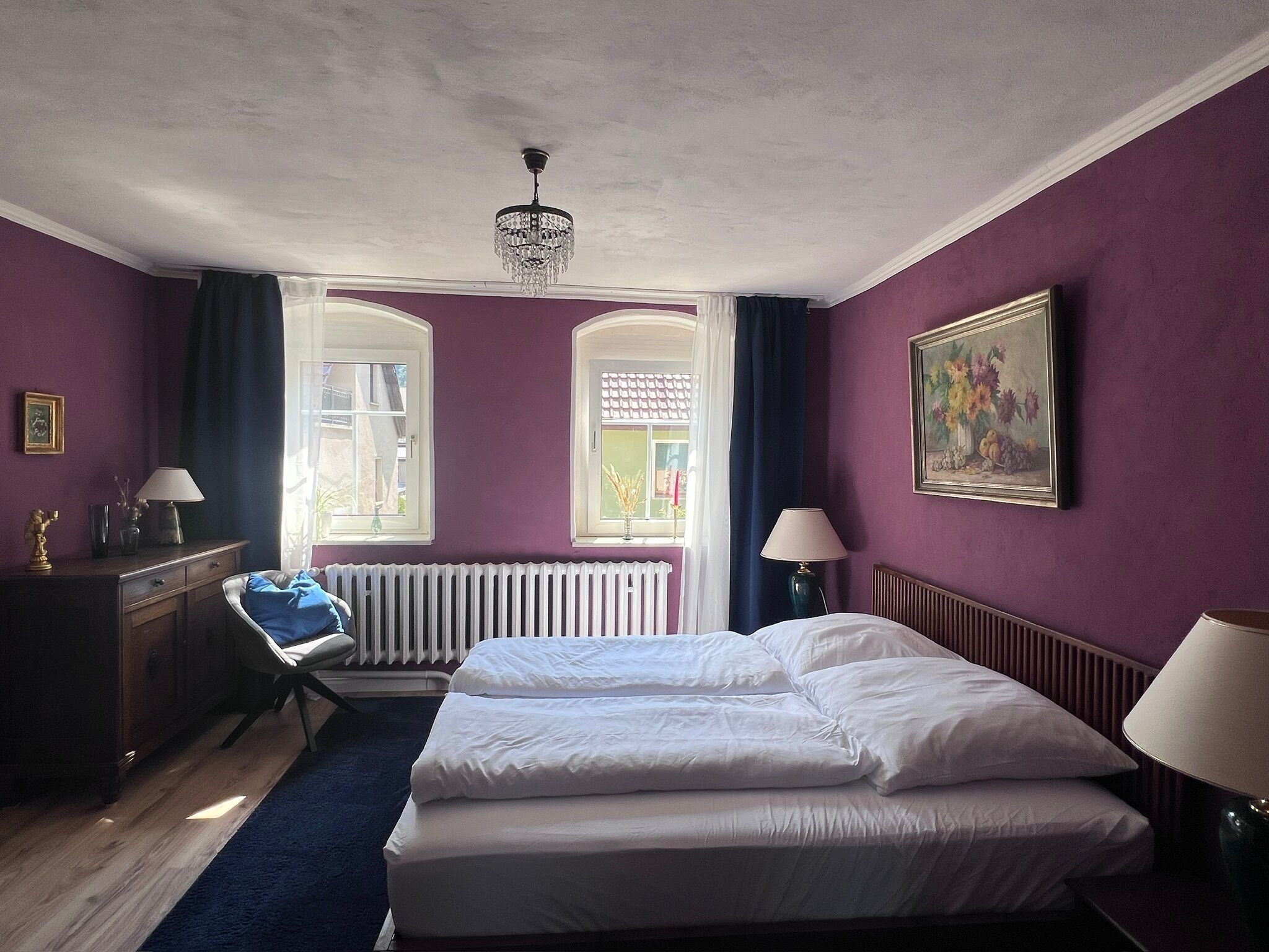 Ein Schlafzimmer mit zwei Betten, einem Heizkörper, einem Schreibtisch mit Stuhl, einem Fenster mit Vorhängen und einer Kristallleuchtre.
