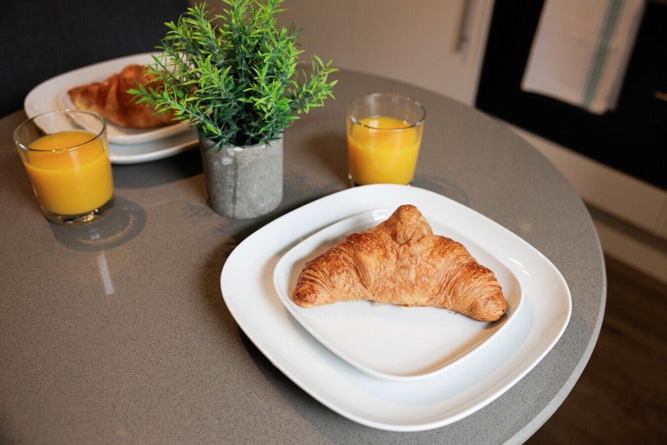 A breakfast setting with a croissant on a plate, two glasses of orange juice, and a small potted plant.