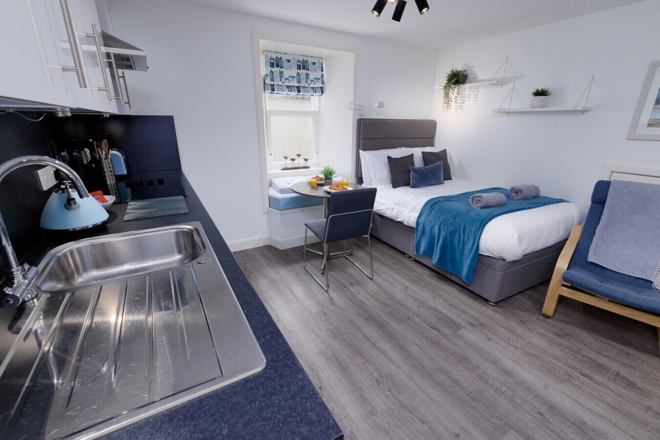 A compact hotel room with a kitchenette, a bed with a blue blanket, a small table, and a chair.