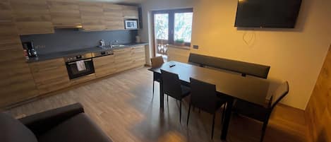 A modern kitchen with a dining area, a flat-screen TV mounted on the wall, and a large window providing natural light.