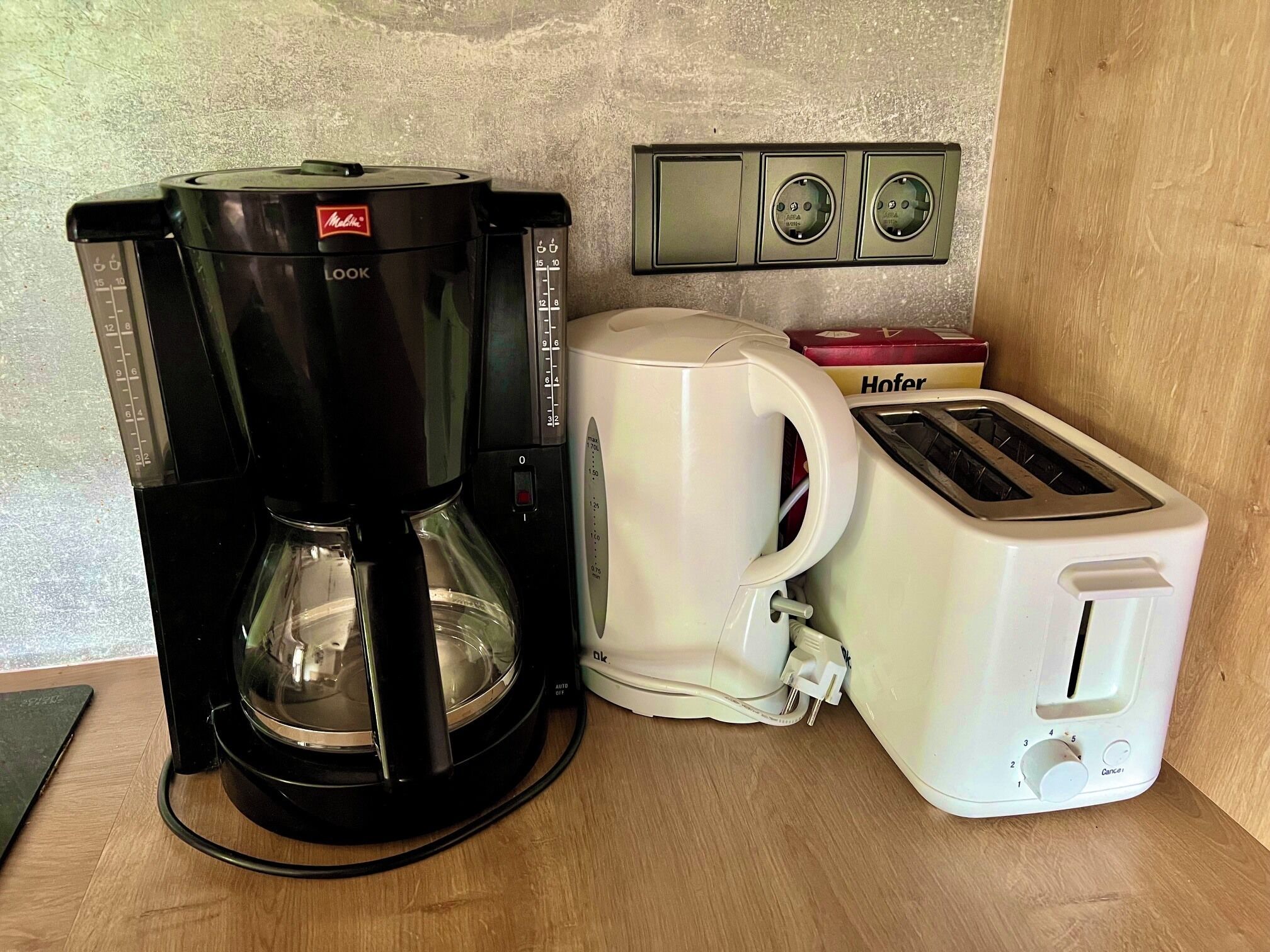 Uma cafeteira, uma torradeira e uma caixa com a etiqueta 'Hofer' sobre uma bancada de madeira.