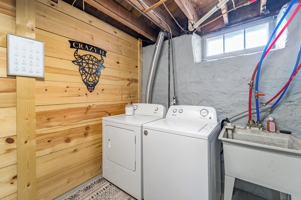 A laundry room with two white washing machines, a wooden wall with a 'CRAZY H' logo, and a window with a view.