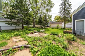 A backyard with a lawn, garden beds, a fire pit, and a patio with chairs.