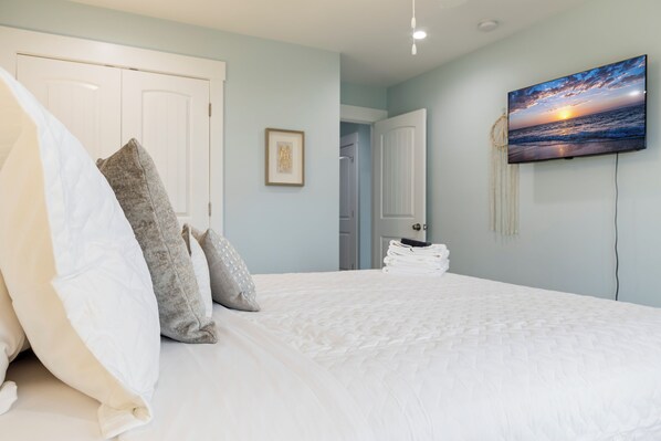 A bedroom with a bed, pillows, a framed picture, a television, and a door. - Ocean Views, Elevator, Fire Pit (Surf City)