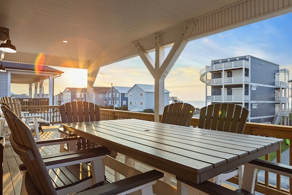 A covered outdoor seating area with a table and chairs, overlooking a residential area with houses and a building with balconies. - Ocean Views, Elevator, Fire Pit (Surf City)