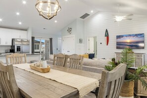 A modern kitchen with stainless steel appliances, a dining area with a wooden table and chairs, and a living room with a sofa and a large painting. - Ocean Views, Elevator, Fire Pit (Surf City)